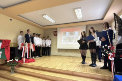 Uczniowie stoją na scenie udekorowanej, kwiatami oraz flagami w kolorach białoczerwonych