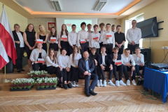 Uczniowie i nauczyciele pozują do zdjęcia, obok stoją białoczerwone flagi