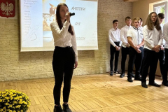 Uczennica stoi z mikrofonem w ręku, w tle inni uczniowie