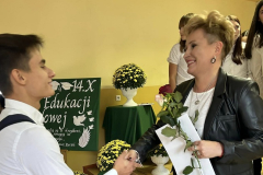 Uczeń wręcza nauczycielce kwiatka, w tle inni uczniowie