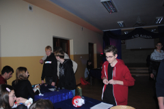 Kilka osób siedzi przy stolikach, obok stoi grupa młodzieży