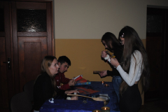 Młodzież siedzi przy stole, obok stoją dwie dziewczyny z kartkami w rękach na stole stoją świeczki