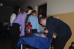 Młodzież stoi przy stoliku, na którym stoi latarenka i leżą kartki