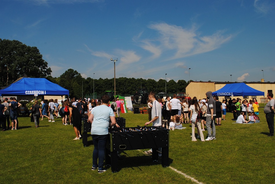 Uczniowie grają w grę piłkarzyki, w tle uczestnicy pikniku, namioty, drzewa