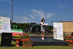 Uczennica stoi na scenie i śpiewa, w tle banery reklamowe, ciągniki