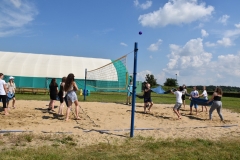Uczestnicy pikniku trzymają ręczniki i podrzucają balon, w tle hala sportowa, niebo