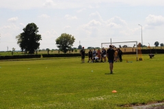 Uczestnicy pikniku podnoszą ciężary, w tle drzewa, bramka, pole