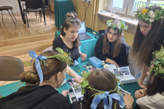 Dziewczęta w wiankach na głowie stoją wokół stołu na którym leżą farby