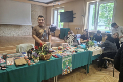 Uczeń stoi obok grupa osób siedzi przy stole na którym leżą fanty