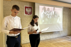 Uczennica i uczeń stoją, w tle godło, zdjęcie z kobietą, meżczyzną i dziećmi