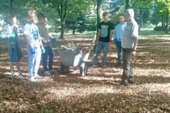 Nauczyciel i uczniowie stoją przy taczce z drewnem, w tle drzewa