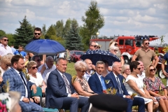 Zdjęcie przedstawia osoby siedzące na krzesłach, w tle drzewa, wóz strażacki
