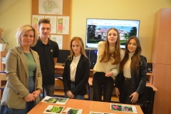 Uczniowie z nauczycielką stoją przy ławce, w tle projekty ogrodów