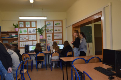 Młodzież siedzi przed komputerami, w tle tablica z rysunkami