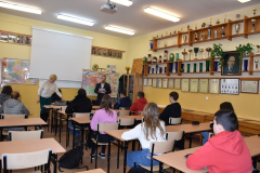 Uczniowie siedzą w ławkach, mężczyzna stoi, w tle puchary szkoły