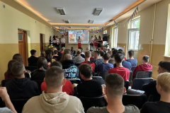 Uczniowie siedzą na krzesłach, na scenie stoi kilka osób