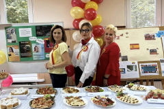 Nauczycielki pozują do zdjęcia stojąc za stołem z potrawami, w tle żółte i czerwone balony
