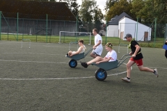 Uczniowie wiozą na taczkach uczniów, w tle budynki, drzewa