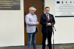 Mężczyźni stoją przy mikrofonie w tle drzwi, napis "Inkubatora Przetwórstwa Owocowo-Warzywnego oraz Wypieku Pieczywa Ziemi Piotrkowskiej"