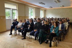 Uczniowie i nauczyciele siedzą na krzesłach, w tle okna