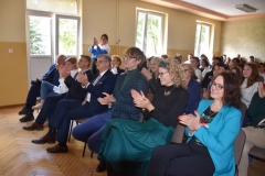 Uczniowie i nauczyciele siedzą na krzesłach, w tle okna