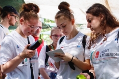 Uczennice stoją przed straganem z papryką i bakłażanami, w rękach trzymają kartki i warzywa
