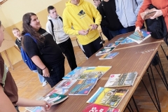 Książki leżą na ławce, obok stoją uczniowie, w tle ściana
