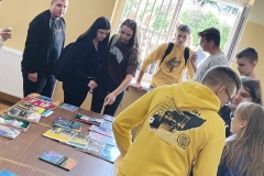 Książki leżą na ławce, obok stoją uczniowie, w tle okno