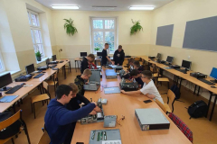 Uczniowie siedzą, na ławce przed nimi leżą komputery, w tle okna, kwiaty, tablice