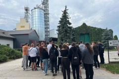 Grupa osób stoi przy budynkach gospodarczych i silosie