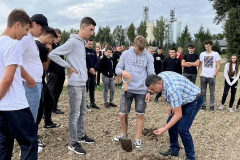 Uczeń trzyma w ręku szpadel, mężczyzna schyla się, w tle stoją uczniowie