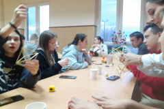 Młodzież siedzi przy stole na którym leżą telefony, stoją papierowe kubki, w ręcach trzymaja bryły geometryczne wykonane ze słomy