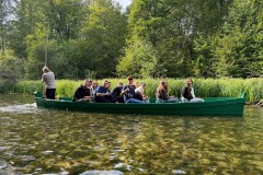 Grupa osób płynie łódką, w tle zarośla, drzewa