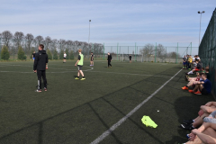 Młodzież gra w piłkę nożną na boisku, przy siatce siedzą kibice, w tle drzewa