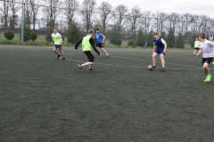 Chłopcy kopią piłkę na boisku, w tle drzewa