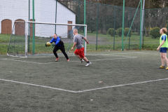 Uczeń stoi w bramce ił łapie piłkę, drugi uczeń biegnie , w tle budynek