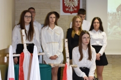 Jedna uczennica siedzi na krześle, pozostali uczniowie siedzą, w tle godło polski