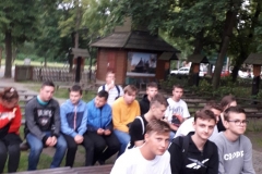 Uczniowie siedzą na ławkach, w tle kapliczka, drzewa