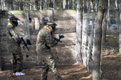 Dwie osoby w strojach moro i kaskach na głowie stoją przy ścianie z desek