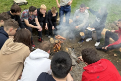 Uczniowie siedzą wokół ogniska i pieką kiełbaski