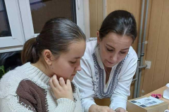 Kobieta w rękach trzyma materiał i igłę z nitką, obok siedzi uczennica, na stole leżą nici, kartki, nożyczki