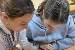 Kobieta w rękach trzyma biały materiał i igłę z nitką, obok siedzi uczennica