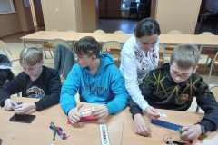 Kobieta stoi, uczniowie siedzą przy stole, na którym leżą kolorowe kawałki papieru, nici telefon