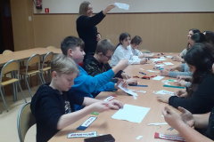 Uczniowie siedzą przy stole na którym leżą nici, kawałki materiałów , nożyczki, nauczycielka stoi w ręku trzyma kawałek białego materiału