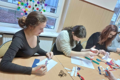 Uczennice siedzą przed nimi na stole leżą wyhaftowane kawałki materiałów, kartki papieru, nożyczki nici