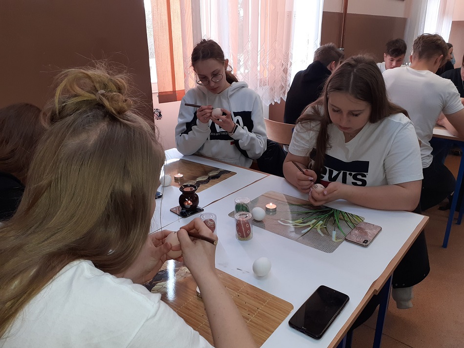 Uczennice siedzą, w jednej ręce trzymają jajko, w drugiej pisak, na ławce stoi rozgrzany wosk, świeczki