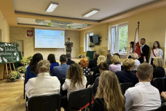 Grupa osób siedzi, w tle stoi mężczyzna