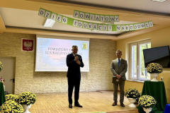 Wicestarosta Powiatu Piotrkowskiego stoi z mikrofonem w ręku, obok stoi dyrektor szkoły, w tle napis Powiatowy Dzień Edukacji Narodowej