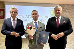Trzech mężczyzn stoi, jeden z nich trzyma w ręku kwiaty i granatową teczkę