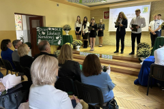 Uczniowie stoją na scenie, grupa osób siedzi, w tle napis Dzień Edukacji Narodowej
